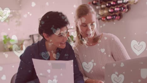 animación de corazones sobre una pareja de mujeres caucásicas sentadas en la mesa y usando una computadora portátil.