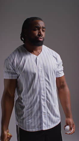 retrato vertical de estudio de video de un jugador de béisbol masculino serio con la camiseta del equipo sosteniendo el bate y lanzando la pelota en el aire disparado contra un fondo gris 1