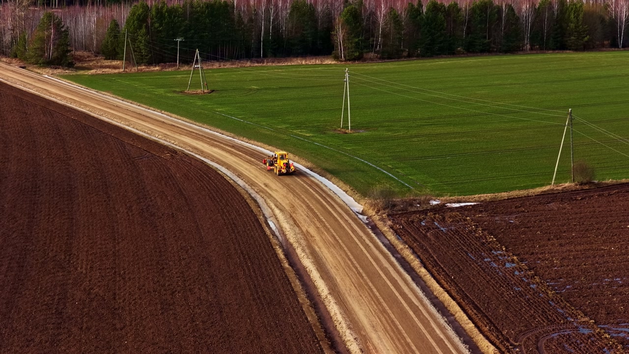 Grader leveling a dirt road, separating a freshly tilled field from green pasture land, aerial view
