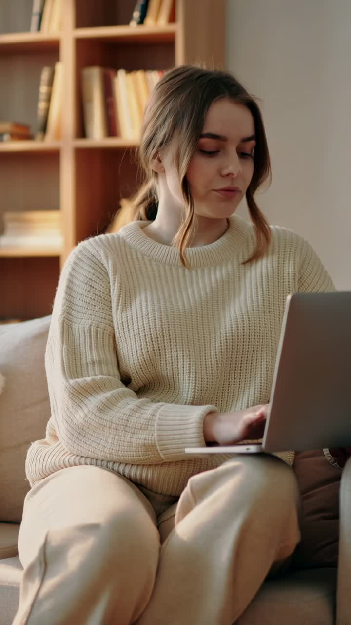 Vídeo vertical: mulher sentada digitando em um laptop no sofá, usando um suéter creme, com uma prateleira atrás