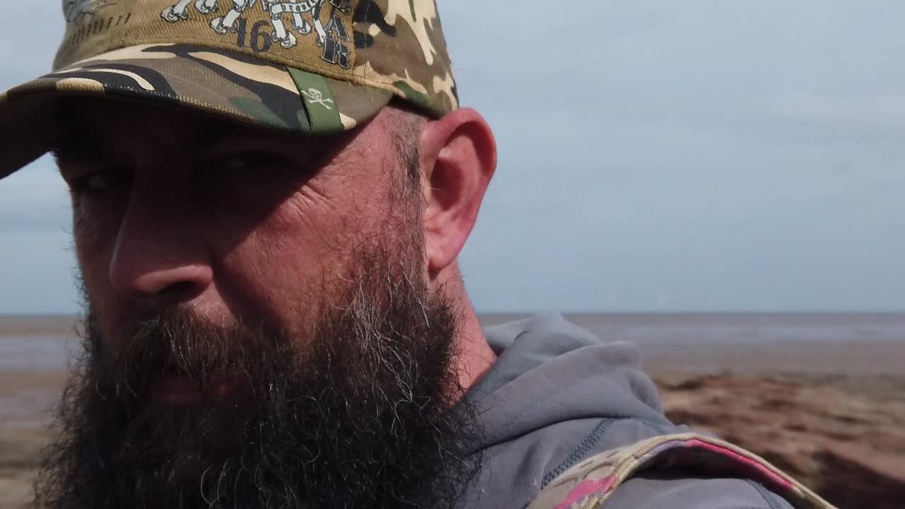 hombre urbano barbudo con gorra de béisbol caminando por la playa ventosa a cámara lenta