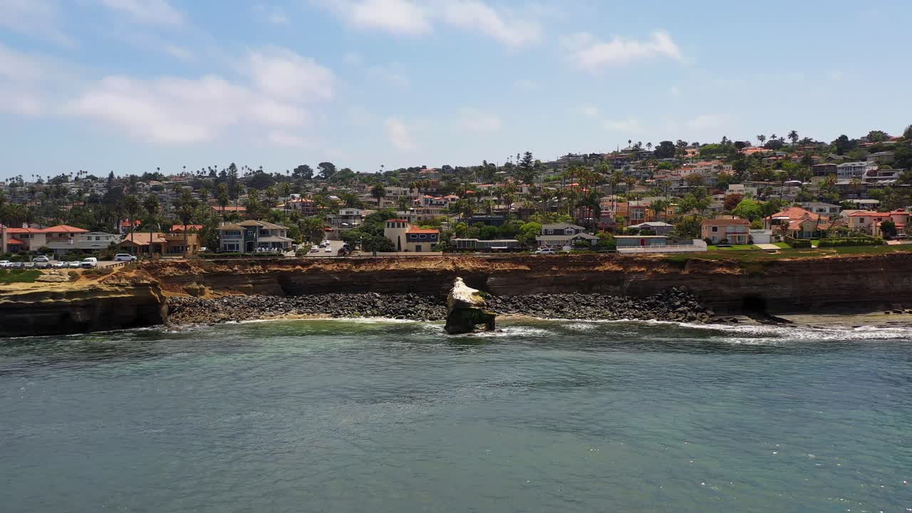 Dron retrocediendo sobre el océano en Sunset Cliffs en Ocean Beach, San Diego, durante el atardecer, con casas costeras y palmeras al fondo.