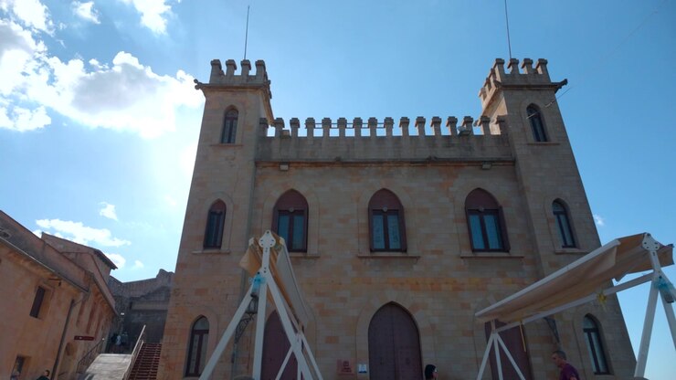 Scena esterna illuminata dal sole che mostra un edificio in stile fortezza in pietra con torri gemelle, finestre ad arco, telai di tettoie bianche, siepi curate e una scalinata accanto a una collina terrazzata sotto un cielo azzurro limpido