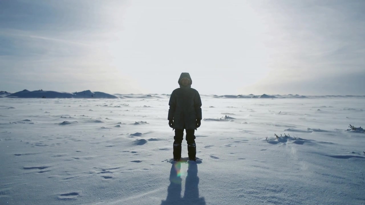 雪原でパーカを着てカメラに向かって立つフードをかぶった人物。太陽の光が明るく、顔は隠れている。
