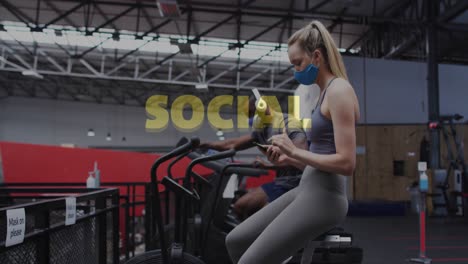 Social-distancing-text-against-caucasian-woman-wearing-face-mask-using-smartphone-at-gym