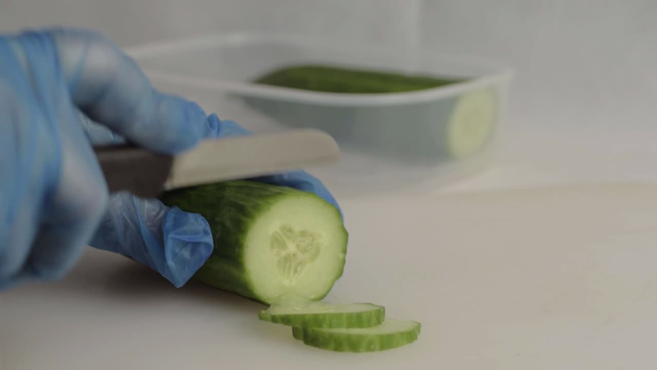 Hand with blue hygiene gloves slicing a cucumber with knife close up