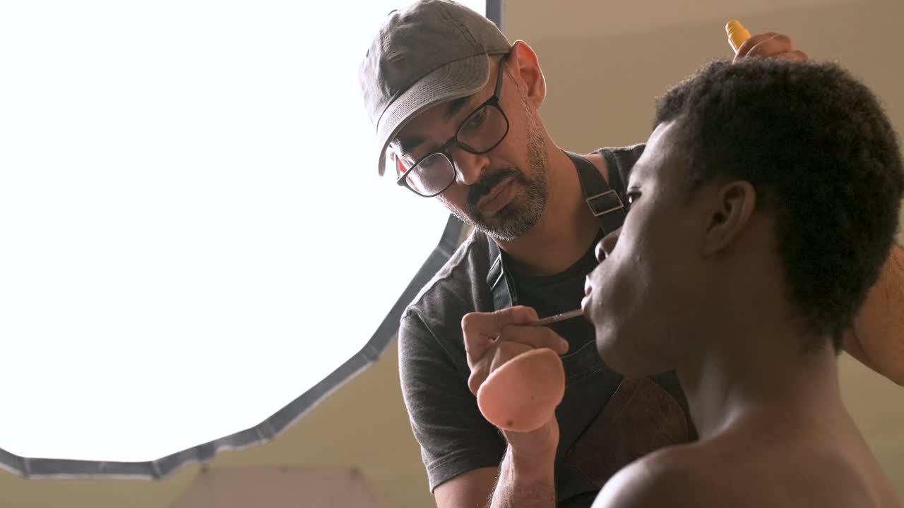 Makeup artist applying lipstick on lips of black transgender in light studio