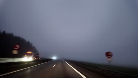 a car is driving on the highway through early morning fog, while a truck and another car are traveling in the opposite direction on the other side of the road.
