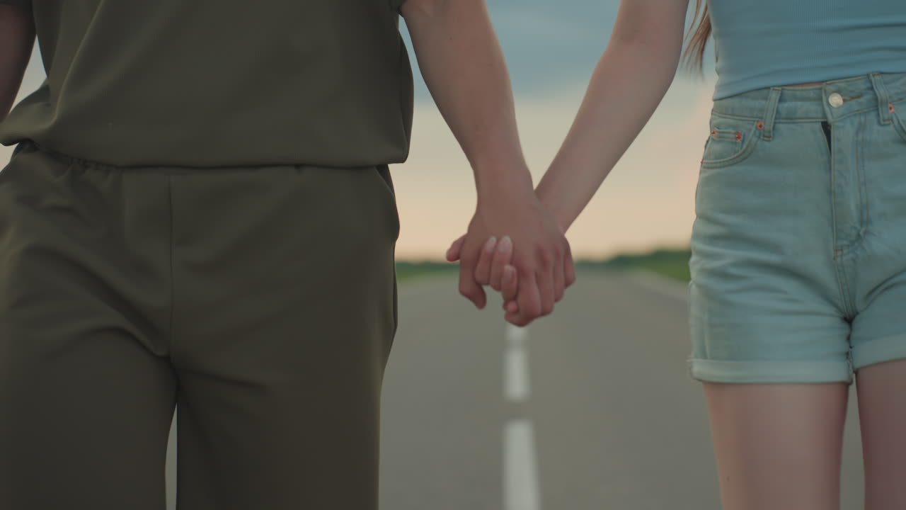 close up of hands of lovers walking side by side on open road at sunset with denim shorts and casual shirt against blurred horizon conveying intimate bond