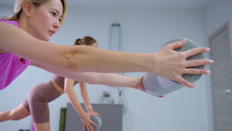 close-up of a woman hand holding a foam roller, extending forward, with another person returning to a normal position and gym equipment in the background