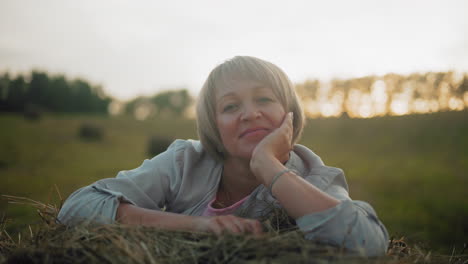 young woman lying on piled hay with warm reflective smile, placing hand under chin, golden sunlight enhances peaceful countryside setting with blurred background