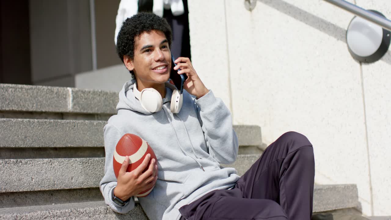 Uomo adulto ispanico che riceve una chiamata, parla al telefono tenendo in braccio un pallone da football sui gradini.