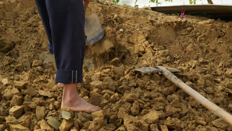 a barefoot man uses a hoe to loosen new clay soil, a raw material for traditional pottery, under the sun, depicting hard labor in rural life