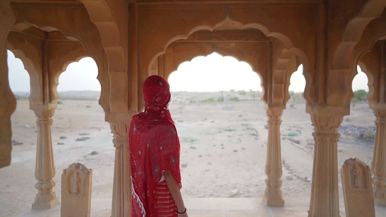A woman in a saree moves slowly and mysteriously through the sandstone cenotaphs of Bada Bagh, Jaisalmer, India, creating an atmospheric and cinematic scene.