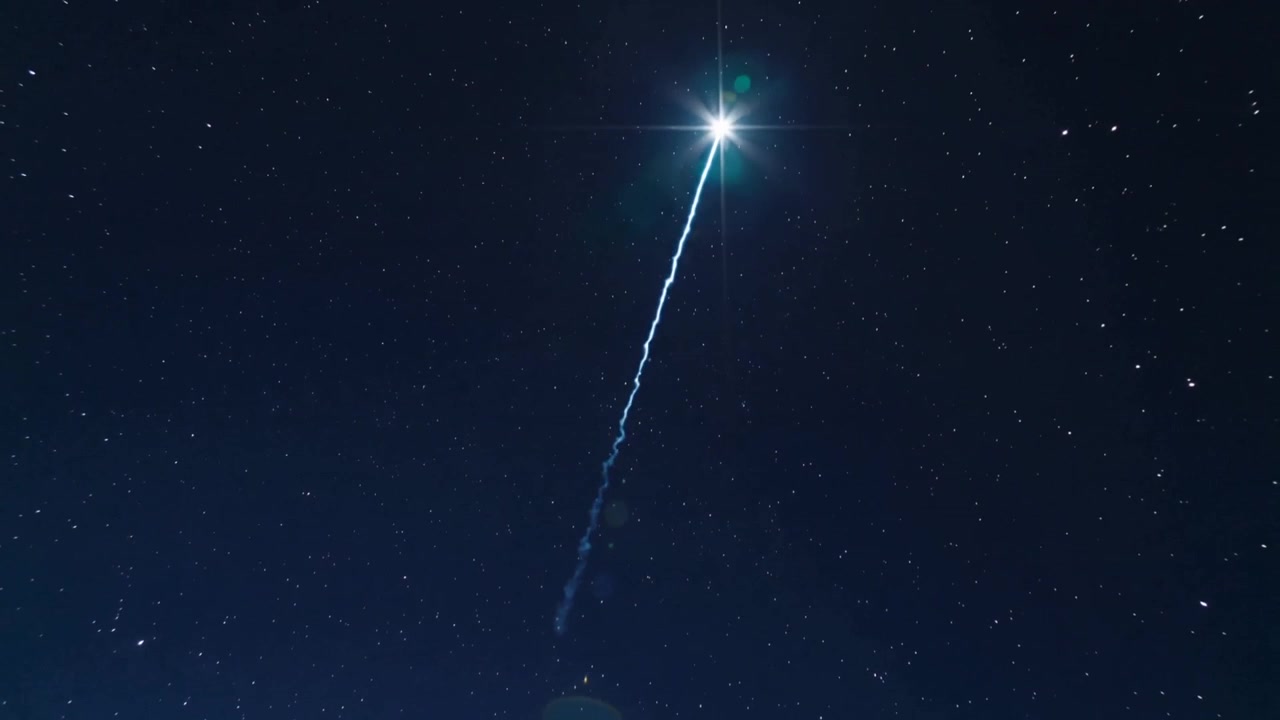 Un bagliore appare, salendo fuori dall'inquadratura nel cielo notturno, lasciando una scia luminosa e lens flare verdi.