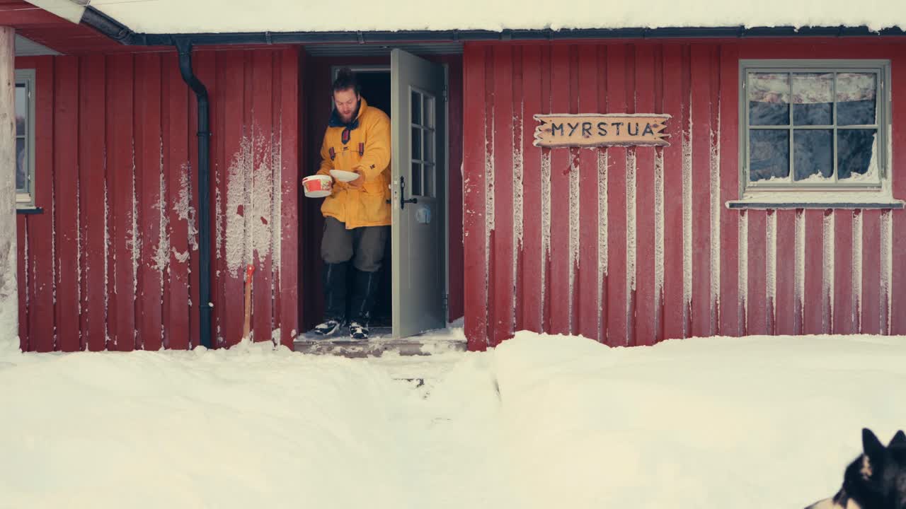 vista frontal de una cabaña de campo con un hombre saliendo por la puerta, trayendo comida para el perro mascota y para él mismo en invierno