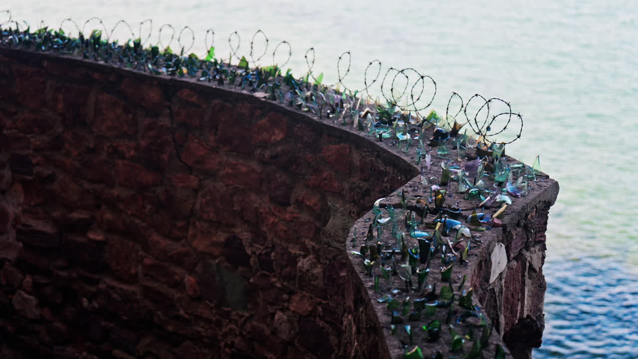 Close up of multiple broken glass shards and barbed wire loops along the edge of a brick wall with a blurred view of the sea