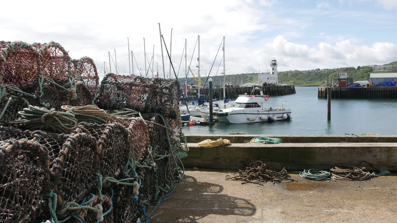 Fishing pots and ropes stacked beside moored boats at the harbor in Scarborough, North Yorkshire in England, with a lighthouse and green hills in the background