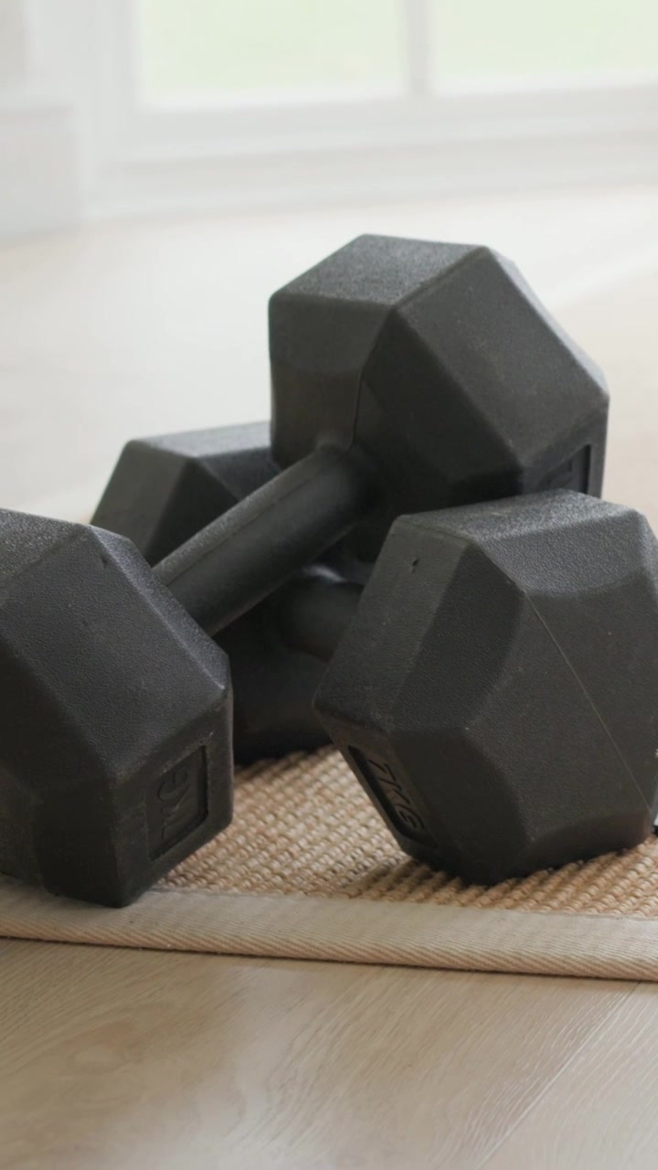 Vertical video: Cam dollying 3 black hex dumbbells on woven mat by window at home, showing home gym