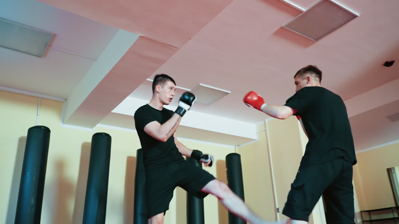 Wrestlers sparring inside gym, wearing gloves and black outfits, practicing combat movements with defensive stance and raised guard on red mat floor surrounded by punching bags