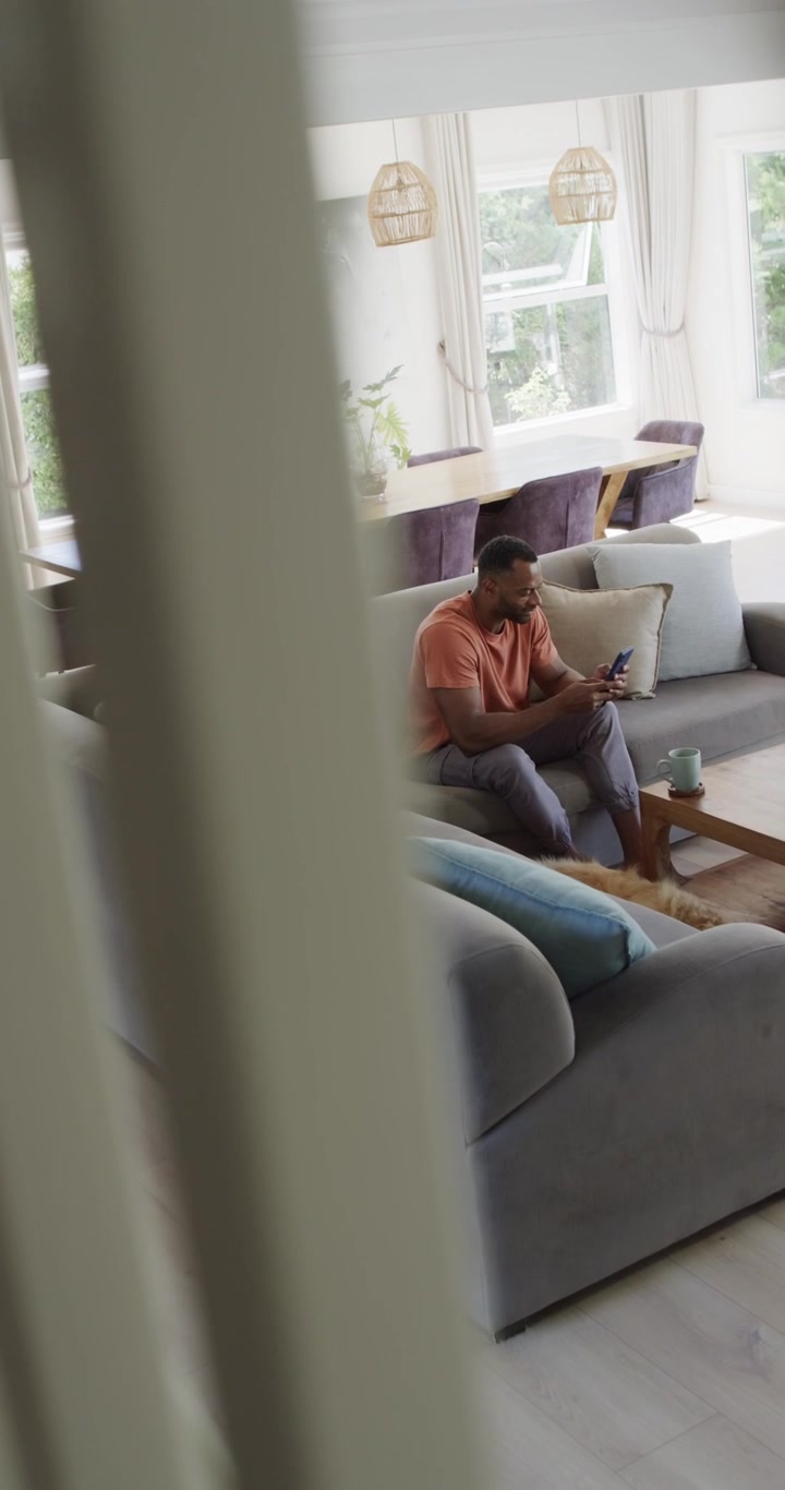 Dikey video: Çıtaların arkasında gri kanepede oturan olgun Afrikalı Amerikalı adam telefona dokunuyor.