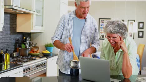 pareja mayor usando computadora portátil mientras cocina en la cocina 4k