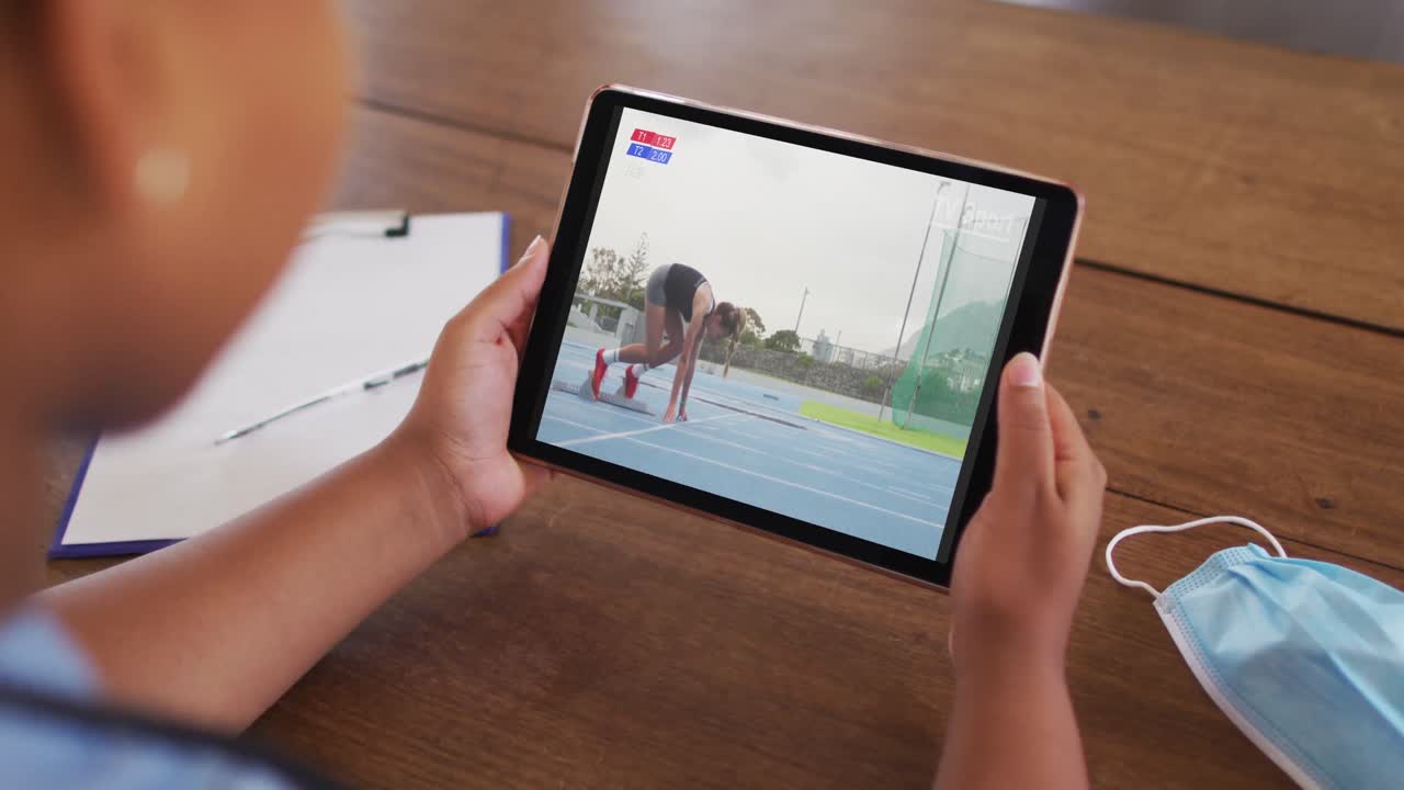 compuesto de una mujer sentada en una mesa con una máscara facial, viendo un evento de atletismo en la tableta