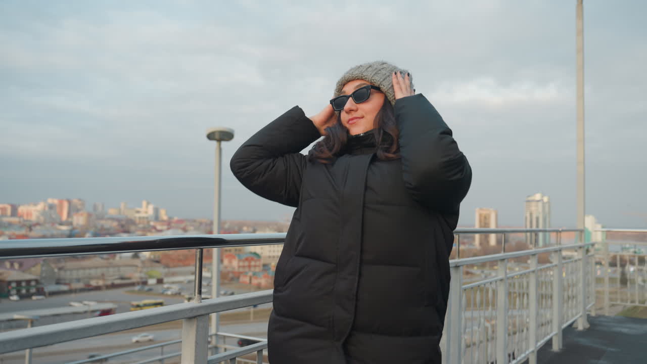 Danza con skyline cittadino: donna con cappotto caldo e berretto grigio si muove con gesti misurati su un balcone sul tetto, isolati cittadini in lontananza, luce cinematografica dell'ora d'oro, si aggiusta gli occhiali da sole.