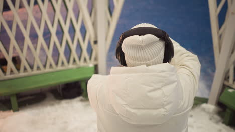 girl wearing white hoodie coat and headphones stands inside snowy gazebo swaying hands gently to unseen music under latticed ceiling with warm lights and snow covered floor visible