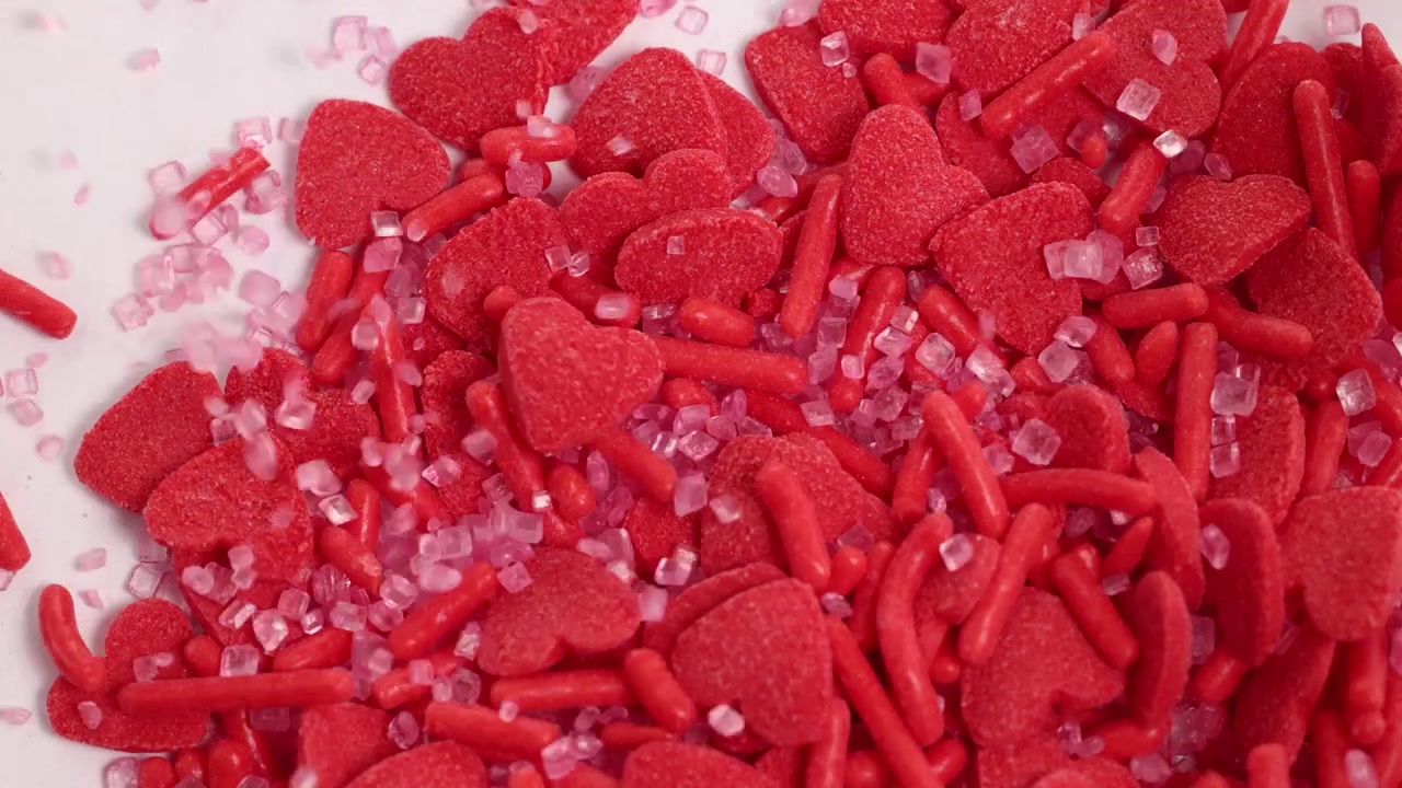 Red heart-shaped candies and sprinkles cascade onto a white surface, creating a vibrant, playful scene with dynamic movement