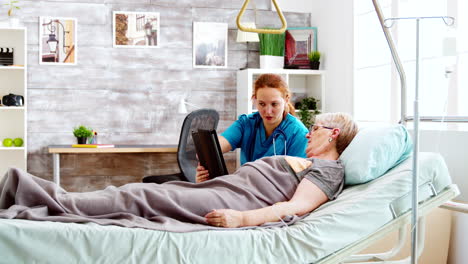 female caregiver helps an old disabled woman lying in hospital bed to use a digital tablet pc