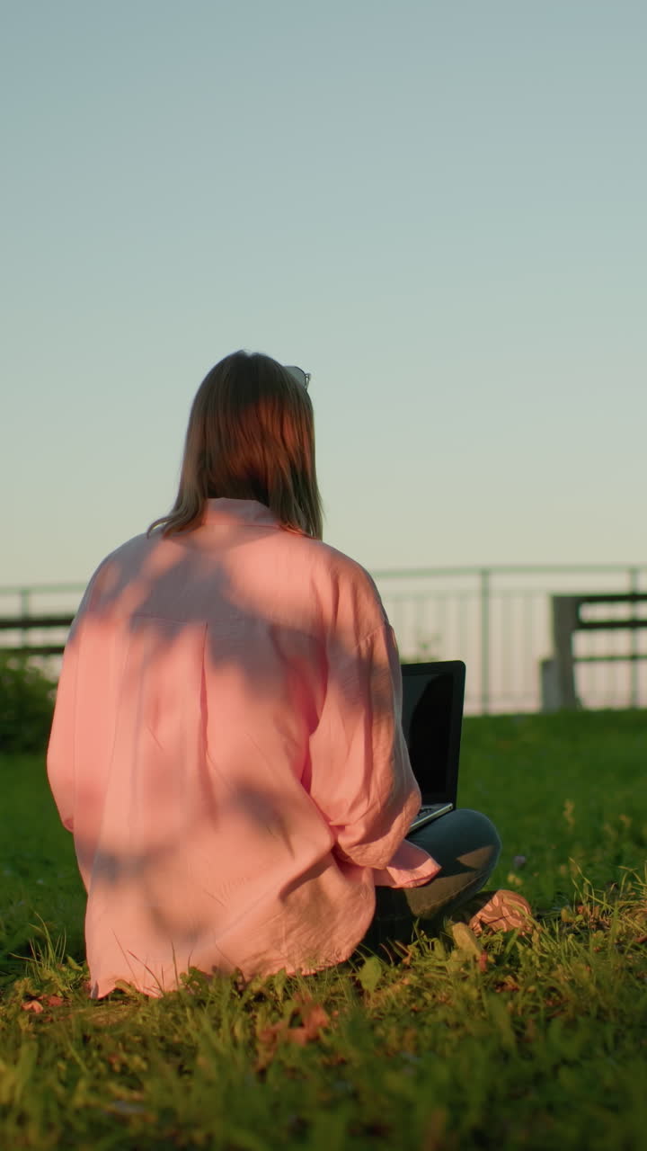 핑크색 을 입은 프리랜서의 뒷면, 안경이 머리에 앉아 있으며, 노트북에 집중하고, 태양 그림자가 옷에 패턴을 만들고, 먼 배경의 벤치