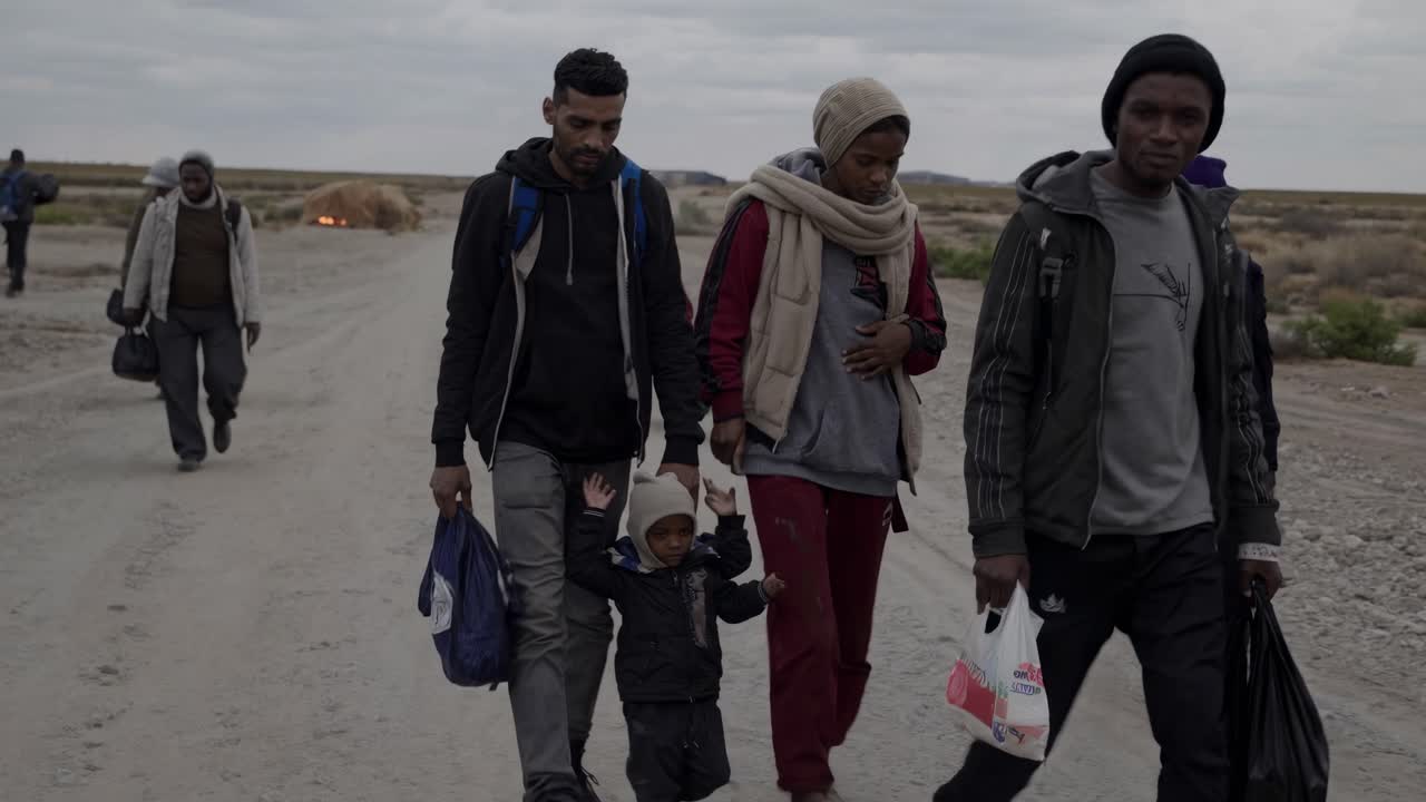 A somber video scene captures a group walking on a dirt road, shot from a low angle