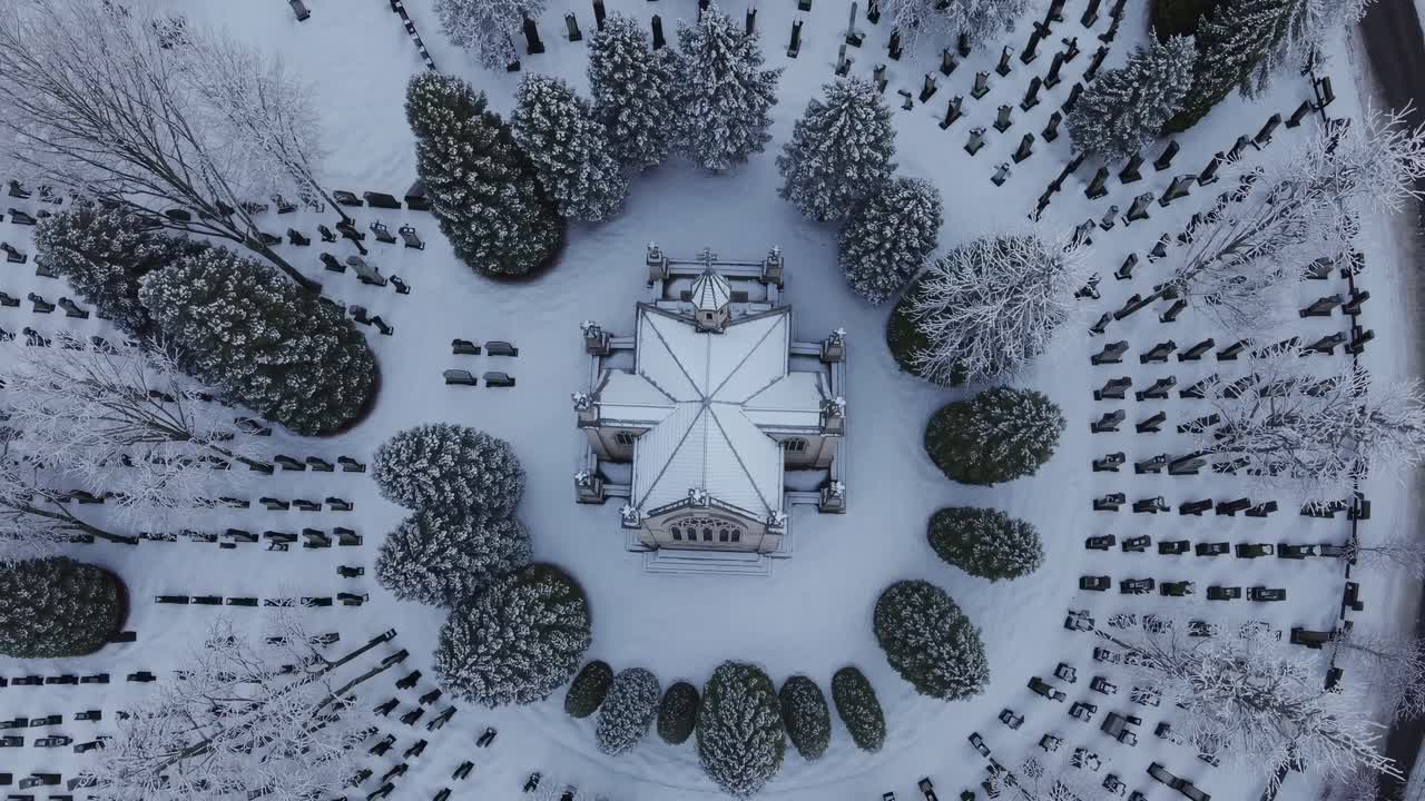 Aerial perspective showcases a snow-blanketed cemetery featuring a central mausoleum, surrounded by lush evergreen trees, evoking a peaceful winter atmosphere