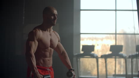 strong purposeful male bodybuilder sport athlete lifting dumbbells holding in hands for training shoulders and back. perform exercises to train your back on the background of the window.