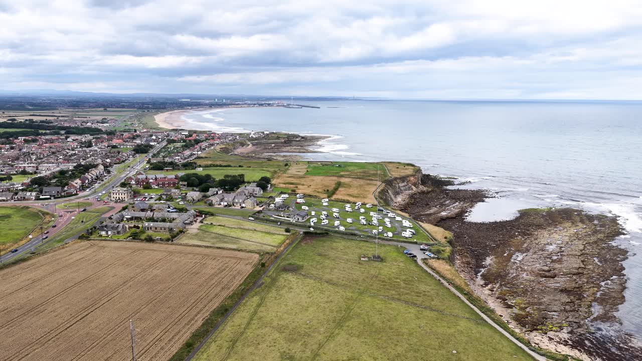 Aerial footage glides over a coastal village, green fields, and rocky shoreline under cloudy skies, revealing houses, farmland, and the expansive sea