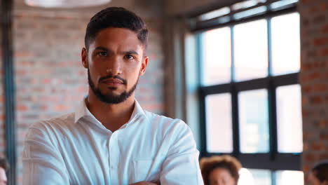 Portrait-Of-Serious-Businessman-Standing-In-Busy-Office-With-Colleagues-Working-In-Background