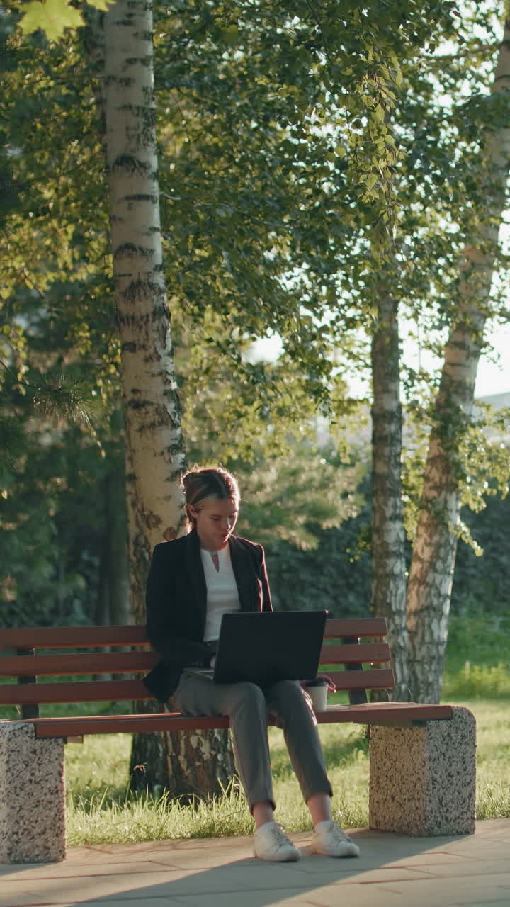 Una lavoratrice da remoto seduta su una panchina di legno all'aperto beve un caffè mentre usa il laptop, circondata da alberi, erba verde, luce del sole e paesaggi urbani