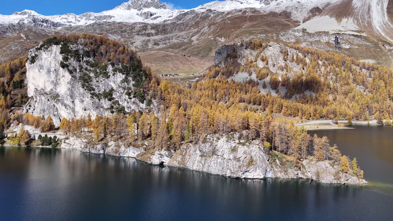 An aerial view of a deep blue Alpine lake, framed by golden larch forests. A small, distinctive rocky peninsula island juts out into the water, backed by snow-dusted mountains