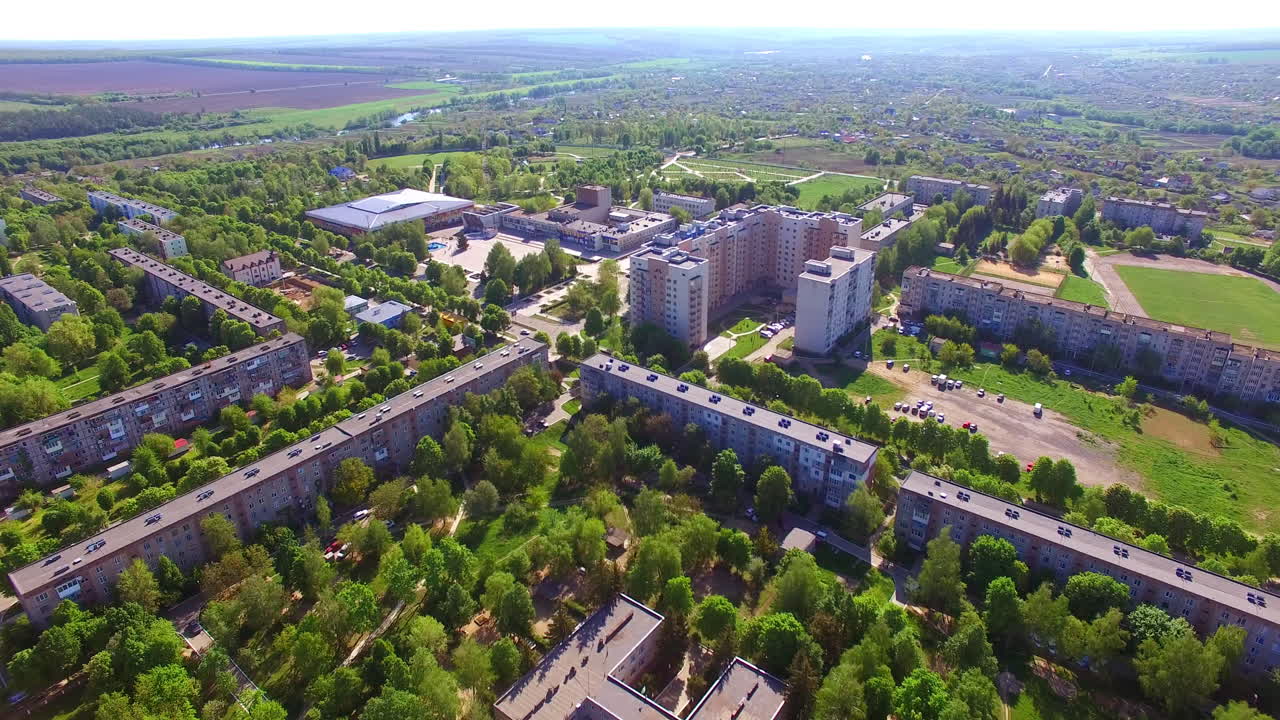 Sunlit panorama of a city with blocks of flats and green streets. Rural territory and farmlands at backdrop.
