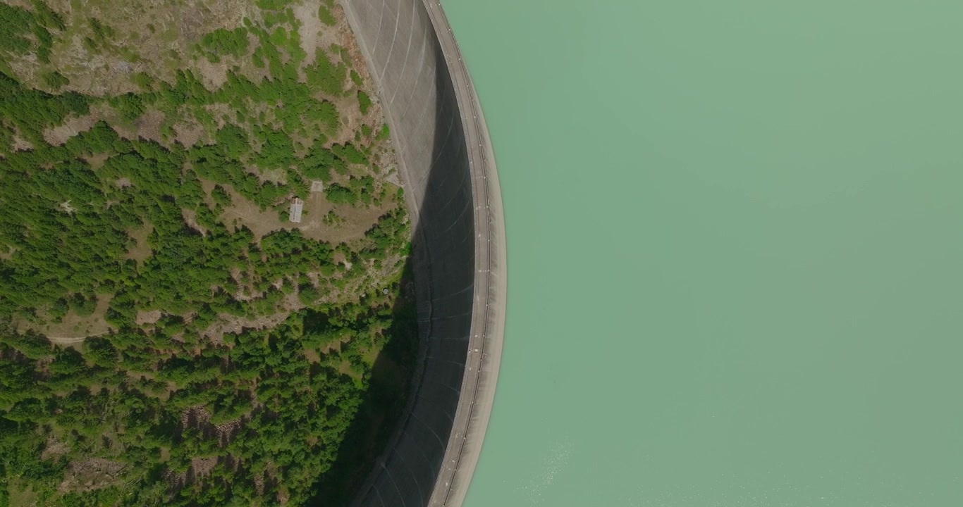 Primo piano aereo con drone dall'alto di un'imponente diga con acqua turchese del bacino su un lato e cime di alberi verdi sottostanti sull'altro, evidenziando l'immensa altezza e la scala ingegneristica.