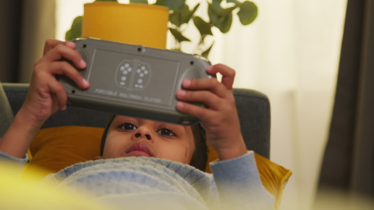 소년은 집에서 소파에 누워서 게임을하거나 휴대용 게임 장치에 스트리밍합니다.