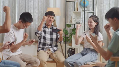 asian teen group sitting in chairs forming a circle playing game on smartphone at home. celebrating success winning game match, giving high five gesture