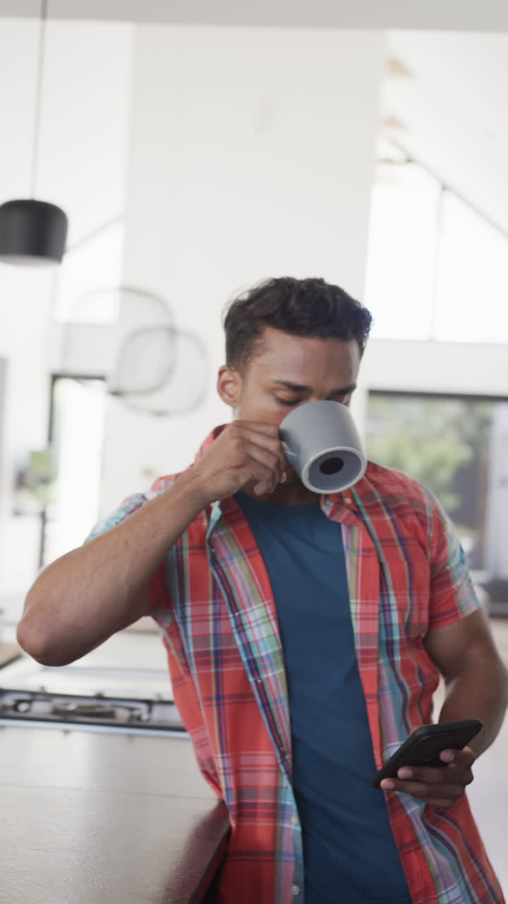 Vertical video of latino man drinking coffee and using smartphone in sunny kitchen