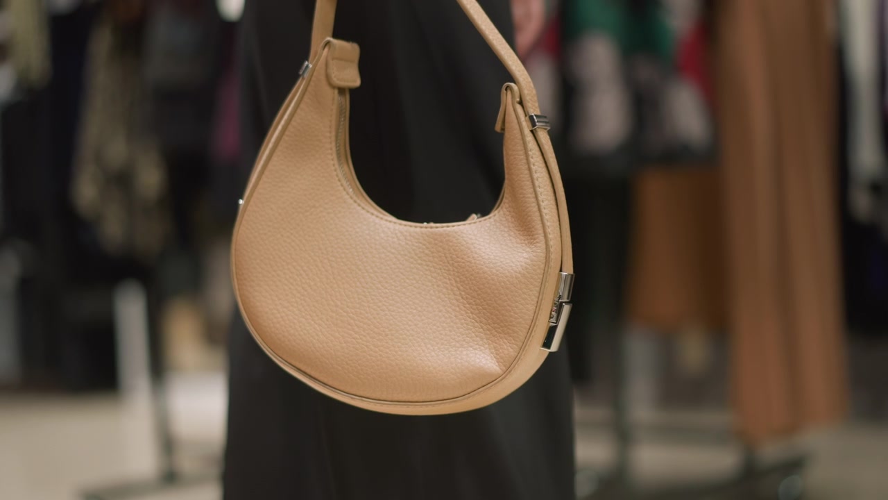 Primo piano di una borsa in pelle beige tenuta da qualcuno in un ambiente di vendita al dettaglio al chiuso. La borsa presenta una texture liscia e dettagli in metallo con un'illuminazione soffusa che ne evidenzia i dettagli su uno sfondo sfocato.
