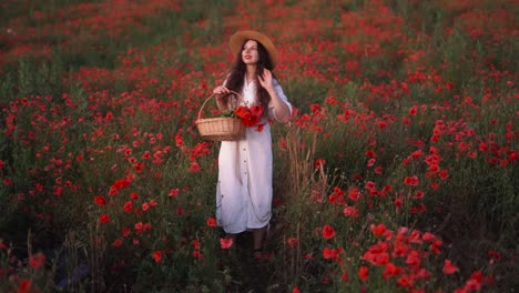 Allgemeiner-Plan:-Schönes-Dunkelhaariges-Mädchen-In-Einem-Feld-Mit-Wildblumen-Und-Roten-Mohnblumen,-Trägt-Einen-Hut-Und-Ein-Kleid,-Hält-Einen-Geflochtenen-Blumenkorb-Und-Lächelt