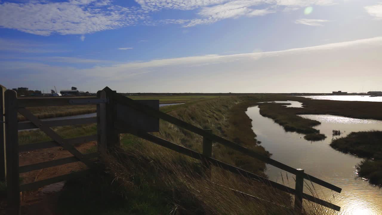 Panning over river and river wall defence with sun reflecting off water and boats in the background. Blue sky with some clouds. Fence and gate in foreground.