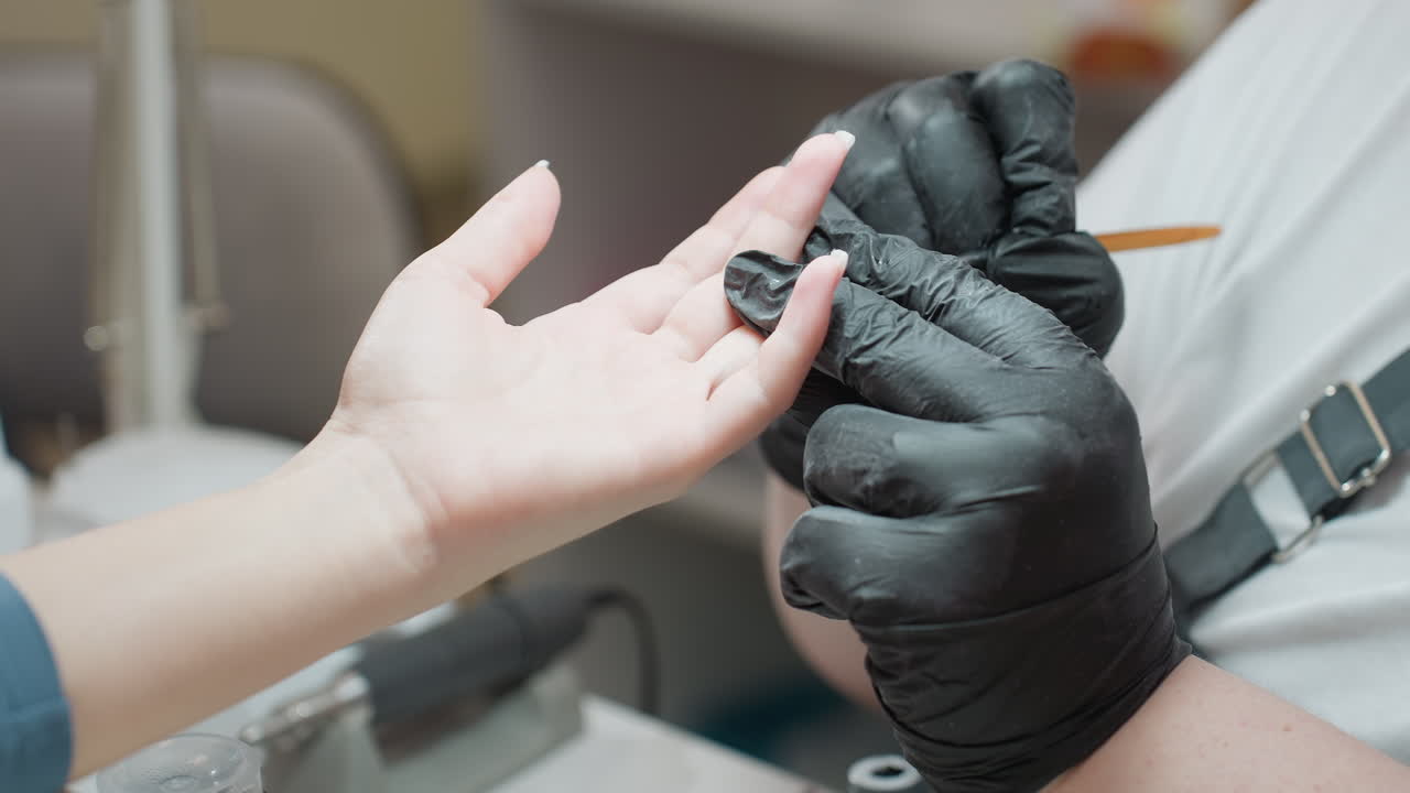 Close-up view of nail technician in black gloves gently applying hardener to customer nail using tiny brush, capturing precision and care in professional manicure service under salon lighting setup