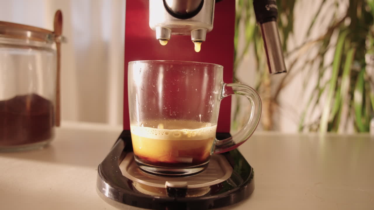 Tomada em câmera lenta com movimento circular de uma cafeteira vermelha servindo espresso fresco em uma xícara de vidro transparente, em uma cozinha caseira aconchegante com luz natural suave da manhã.