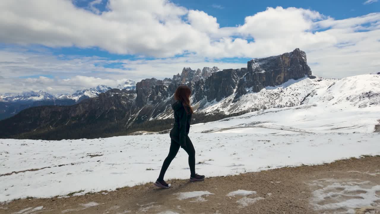 A slow-motion video of a girl walking in Passo Giau, Italian Dolomites, with snow-covered mountains under a cloudy sky